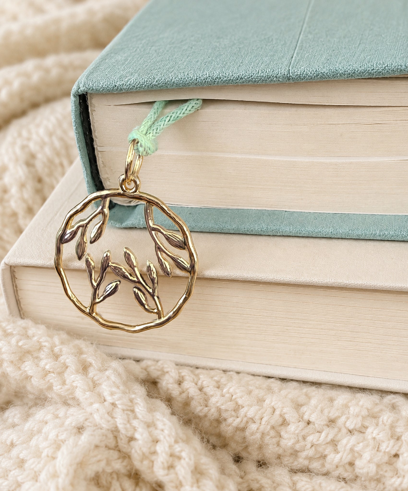 Round gold blossom charm on light green cord hangs from a book stack on cozy blanket.
