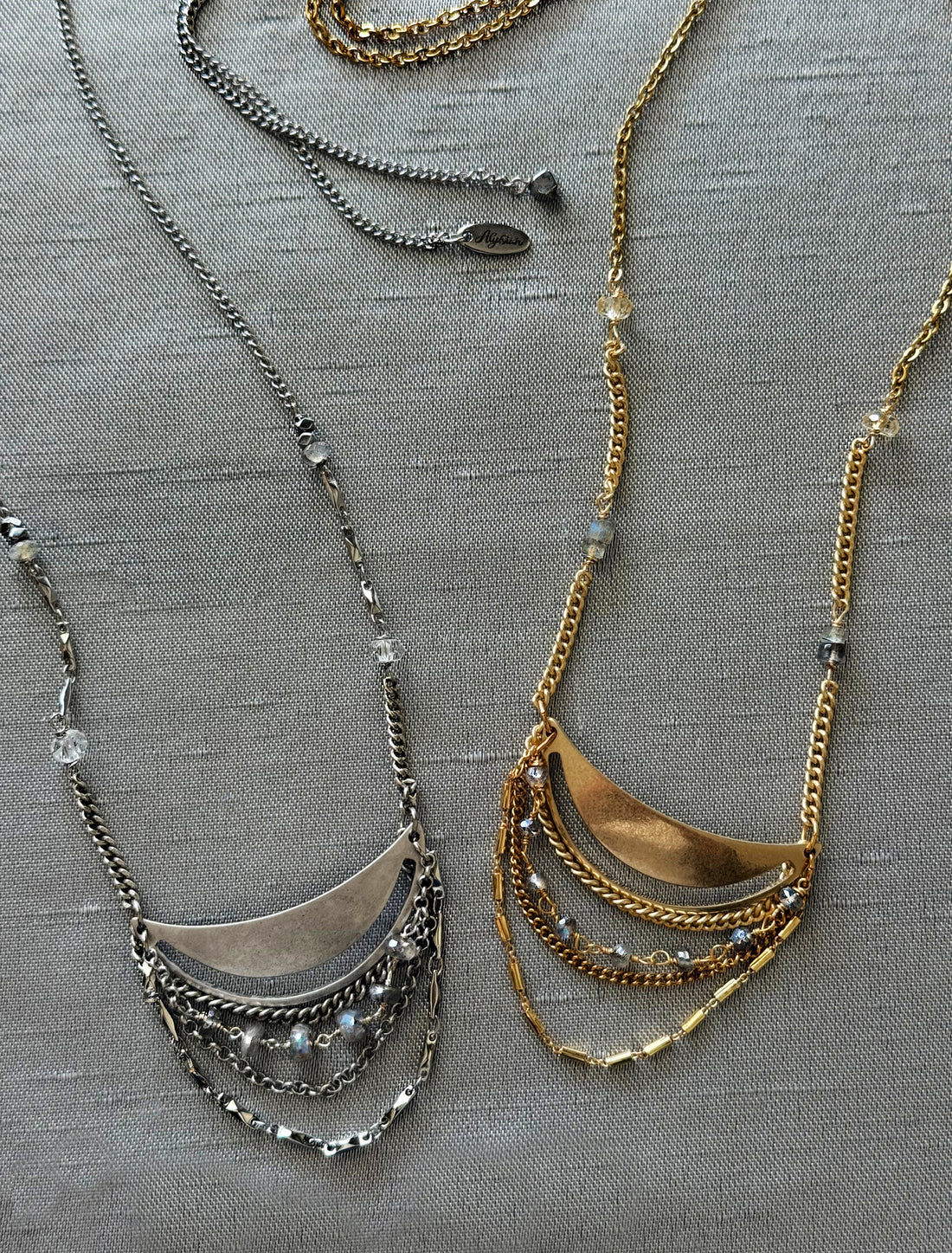 Two Lusine adjustable crescent pendant necklaces with draped chains and stones, one silver and one gold, on a luxe, textural fabric background.