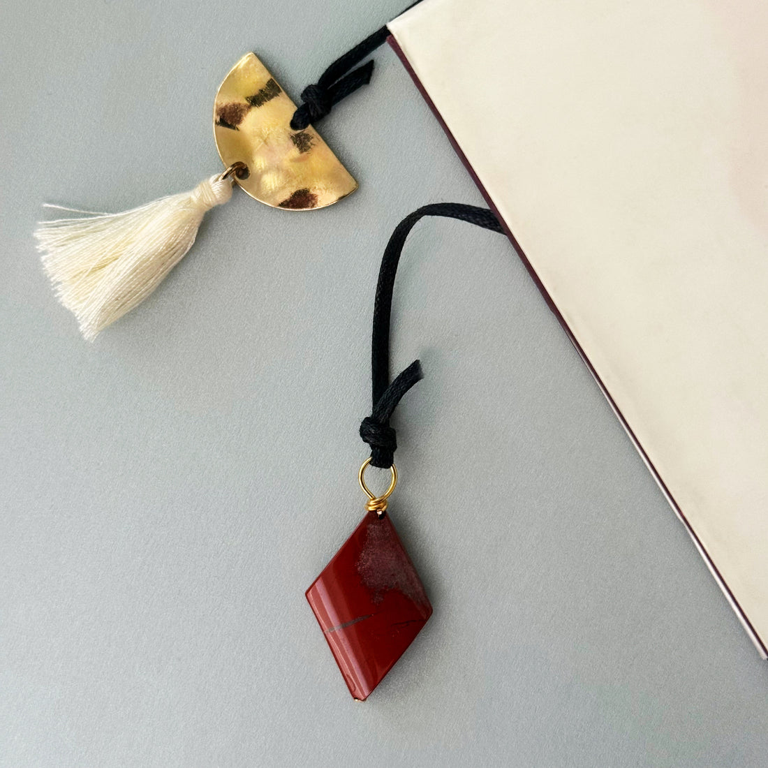 Decorative Alysian Bria Red Jasper Bookmark with red stone and gold accent and cream tassel with a book on a light gray surface.
