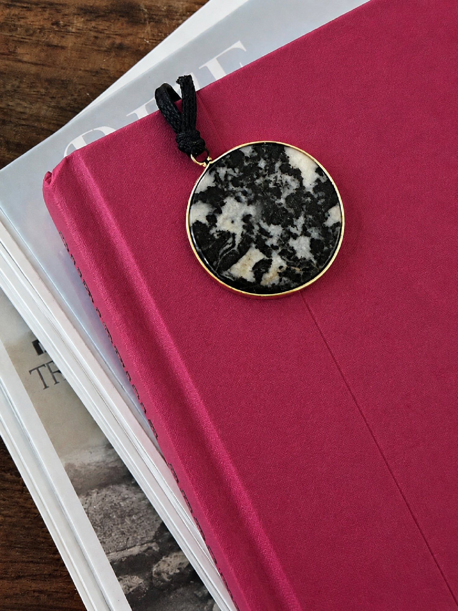 Round and flat black and white zebra jasper pendant on a black cord, peeking out of deep pink book.