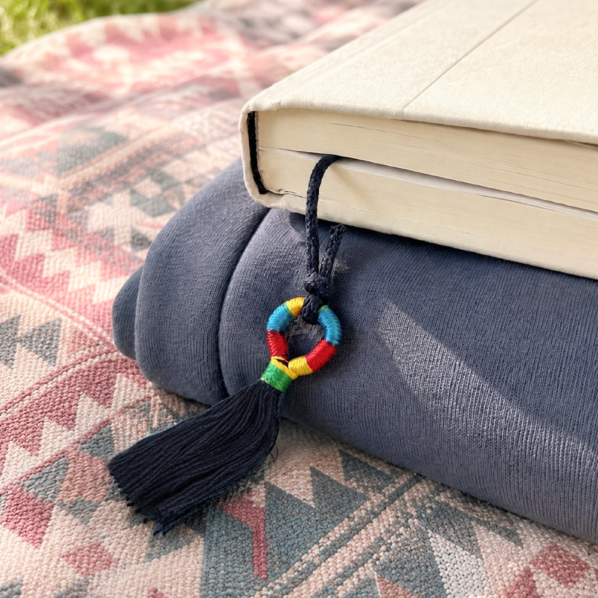 Colorful tassel Inanna Bookmark peeks out of book on top of a folded sweatshirt and a textural  boho picnic blanket outdoors.