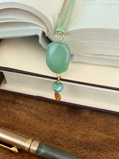Green aventurine stone bookmark dangles from an open book with a pen next to it.