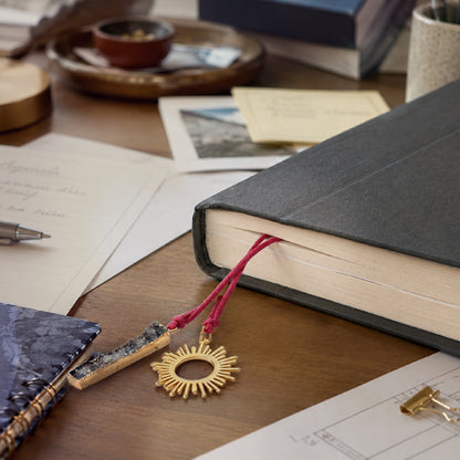 Nova bookmark with a gold sun-shaped pendant and rectangle gray druzy stone on fuschia cotton cord from  a book with stationery items on a wooden desk.