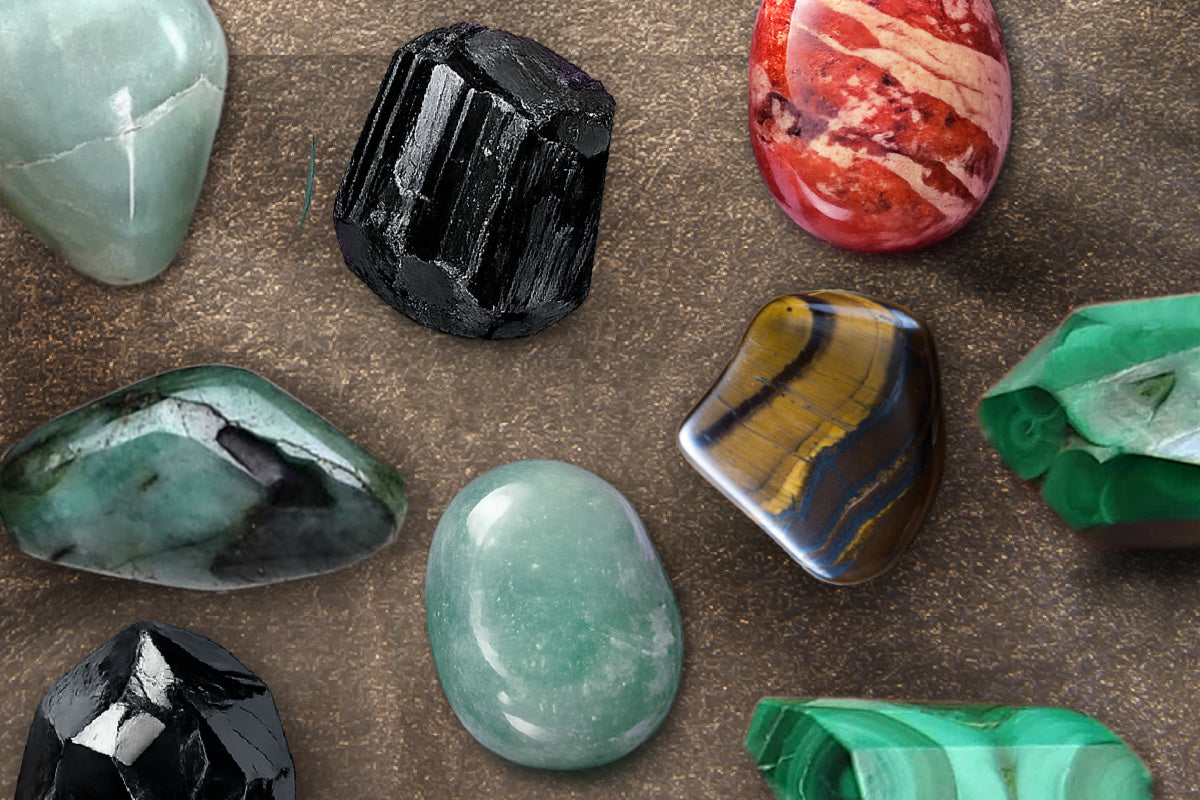 An array of various gemstones including red jasper, black tourmaline, obsidian, jade, malachite, emerald and tiger's eye. All smooth and faceted stones are polished and appear on a textured surface.