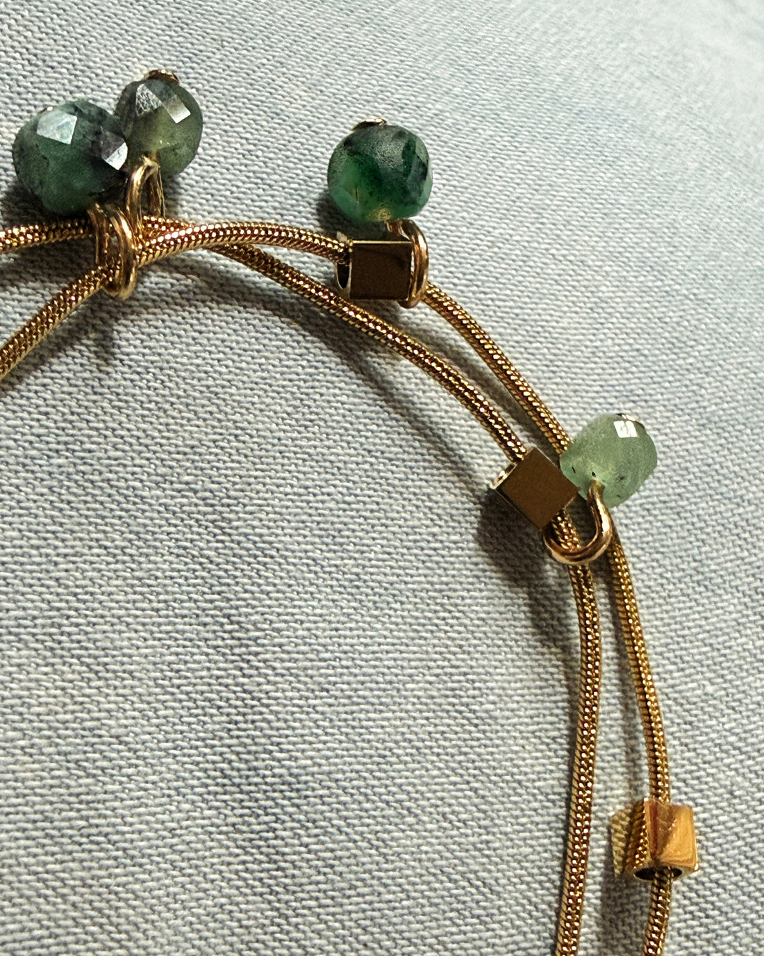 Modern layered gold tone bracelet with green emerald stone beads shimmering against a fabric background. Handcrafted by Alysian.