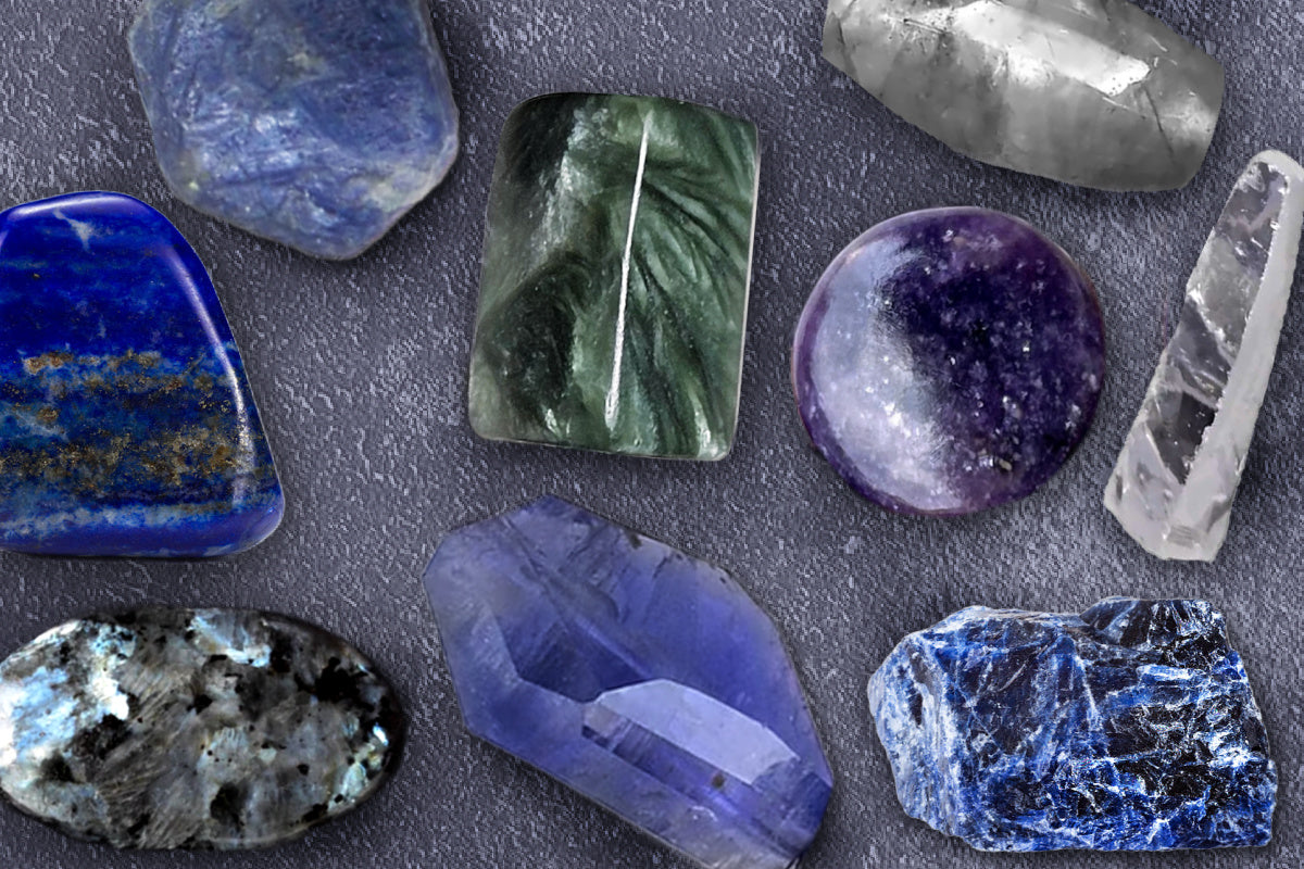 Collection of various crystals and stones with a moody and cool vibe, including blue sapphire, lapis lazuli, larvikite, iolite, zircon, cloud quartz, seraphinite and lepidolite against a dark surface.